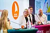 Paneldeltakere på en konferansescene med mikrofoner og flasker foran en bakgrunn med logo.